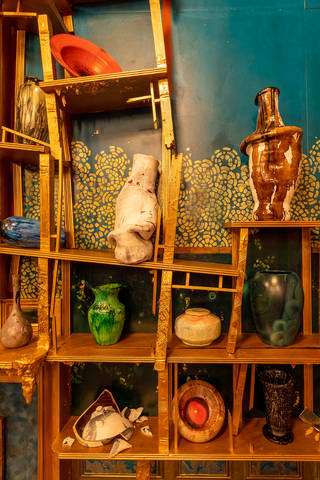 Broken slanted shelves holding colourful ceramic pots, plates and vases, some of which are is broken