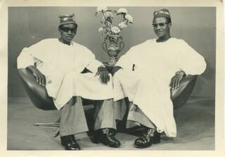 Two men wearing white shirts, hats and sunglasses, sitting back in chairs. There is a vase with flowers behind