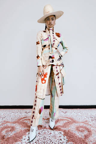 Woman wearing a cream suit with patchwork detailing, a large straw hat tied under the chin, and white pointed heeled boots