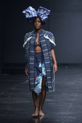 Person on catwalk wearing headpiece, jacket and skirt made from dyed cloth in shades of light blue, dark blue and white