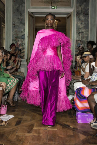 Woman on catwalk wearing metallic pink trousers, top and cape with tasselled detailing