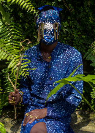 Person wearing dark and light blue buttoned dress with speckled design, and blue mask with beaded detailing
