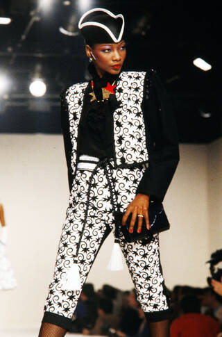 Woman on the catwalk wears black and white waistcoat trouser set and pirate-style hat. She looks to her left.