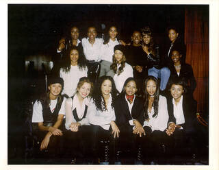 Black female models stood/sat in three rows for a group photograph. They are all smiling. 