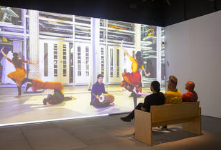 Three people sit in a gallery space watching a projected film of dancers